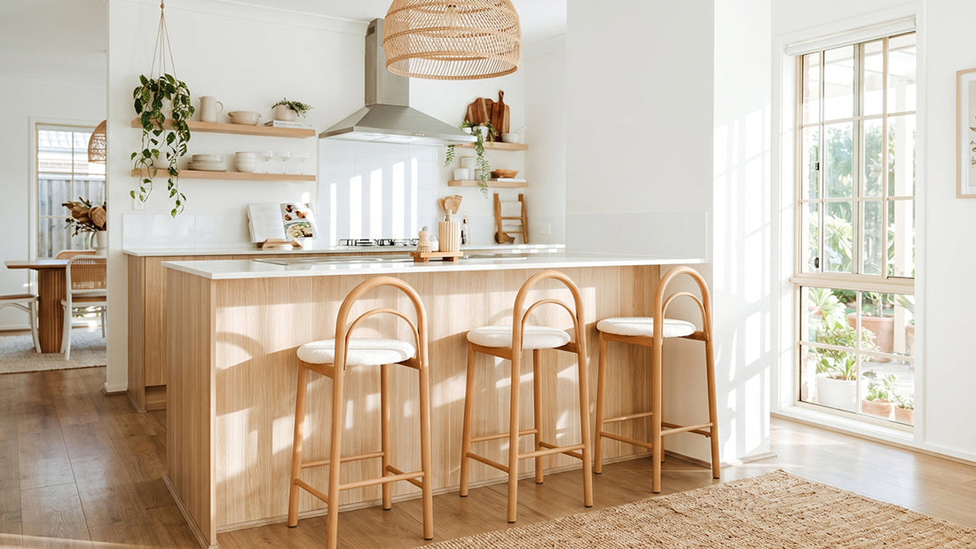 A bright, modern kitchen featuring a light-timber kitchen island with a white countertop, paired with three matching bar stools that have light-toned wooden frames, arched backrests, and round, off-white upholstered seats, situated on a woven rug