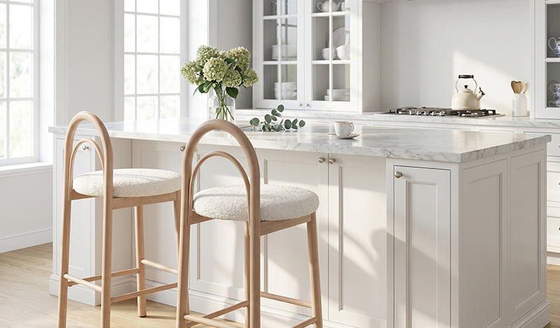 A bright, airy kitchen featuring a white island with a marble countertop, paired with two bar stools that have light-toned wooden frames, distinct arched backrests, and round, off-white upholstered seats.