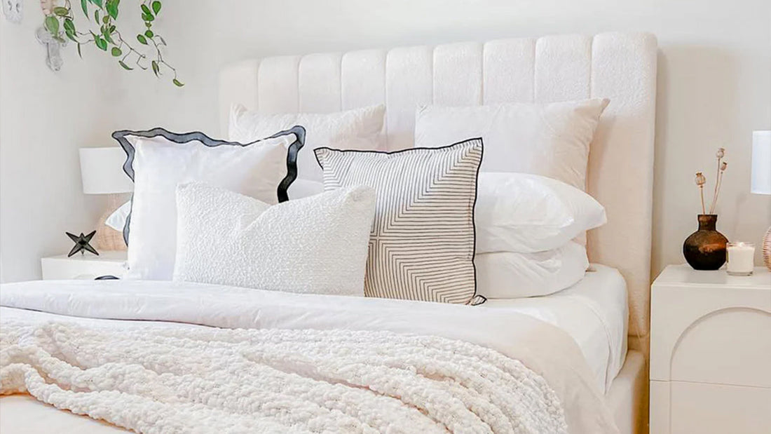 Close-up of a plush bed with a tall, light-pink upholstered headboard and layers of white and patterned decorative pillows and blankets.
