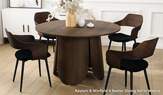 A modern four-seater dining set in a rich walnut finish, featuring a round table with a distinctive, curved-panel pedestal base and four matching dining chairs with walnut-veneered backrests, black upholstered seats, and slim black metal legs.