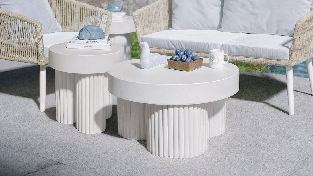 Basalt & Salvo Outdoor Concrete Coffee Table Set in White features two round tables with fluted bases, adorned with a marble bird sculpture, plums in a wooden bowl, and a patterned ceramic mug.