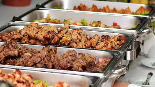 Commercial kitchen buffet setup with stainless steel chafing dishes and grilling tools serving skewered grilled meats and vegetables.