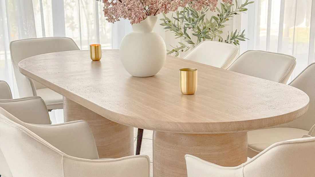 Oval light wood dining table on a sculpted base, surrounded by cream-colored dining chairs, topped with a white vase and two gold candle holders.
