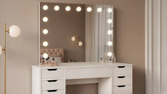 White dressing table with multiple drawers and a large vanity mirror framed by bright circular light bulbs, set against a neutral-toned wall.