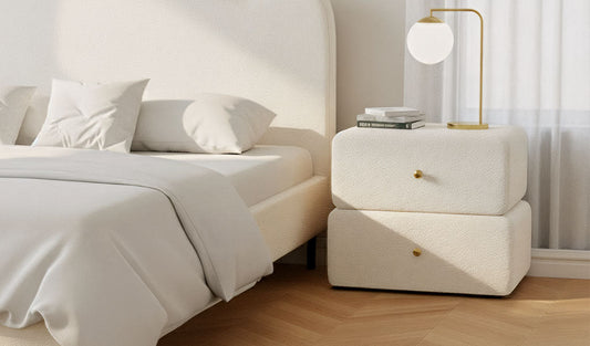 A modern bedroom features an off-white upholstered bed and a chunky, two-drawer bedside table covered in a cream textured fabric (like boucle) with small brass pulls. On top is a simple brass lamp with a spherical white shade.