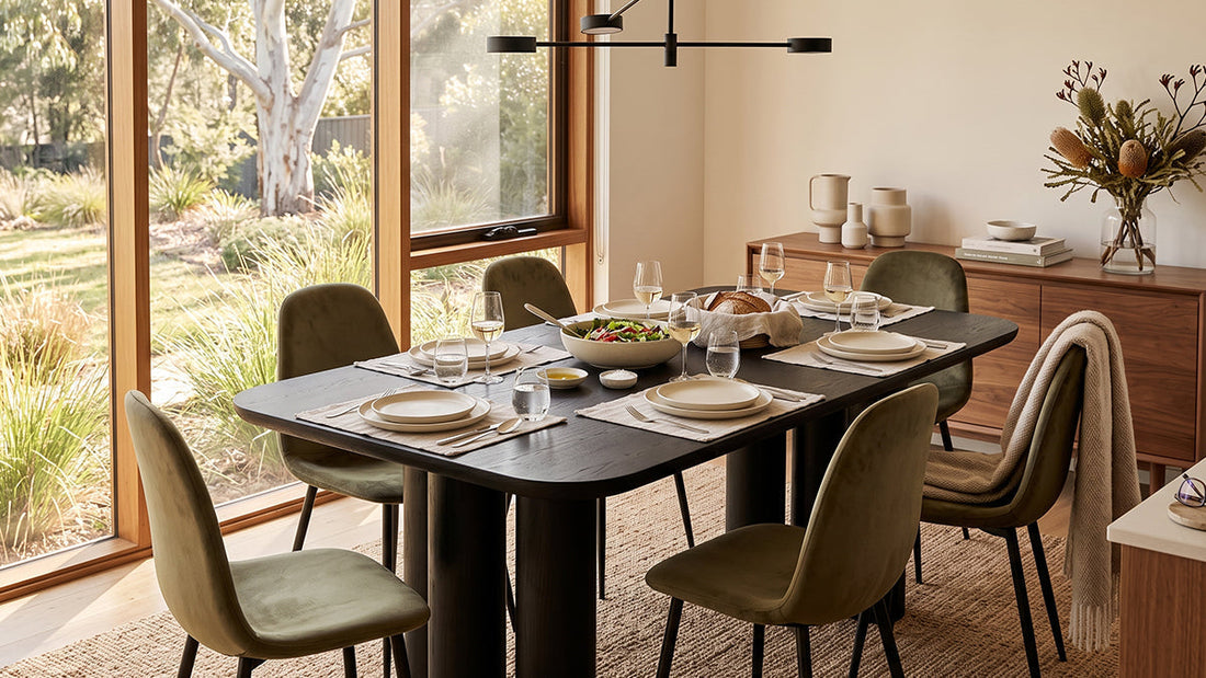 A modern dining room setup featuring a dark-toned, rounded-corner table surrounded by olive-green velvet upholstered dining chairs, with a large window and wooden sideboard in the background.