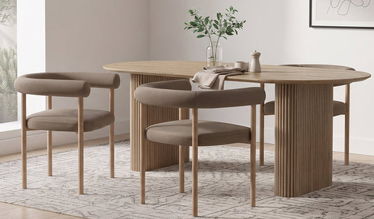 A modern dining setup featuring two dining chairs with light-oak frames and taupe-coloured upholstered seats with curved backrests, positioned beside an oval dining table with a matching light-oak, fluted pedestal base on a textured rug.