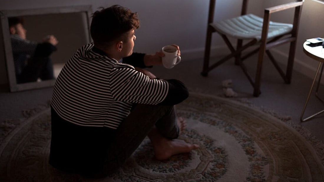 A young person with short, dark, curly hair is sitting cross-legged on a patterned round rug in a dimly lit room, holding a white mug.