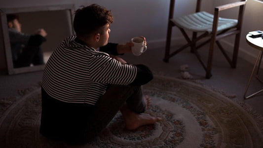 A young person with short, dark, curly hair is sitting cross-legged on a patterned round rug in a dimly lit room, holding a white mug.