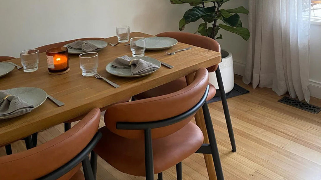Dining table set for four with a light wood top, leather-upholstered chairs, a candle, and plates with napkins.