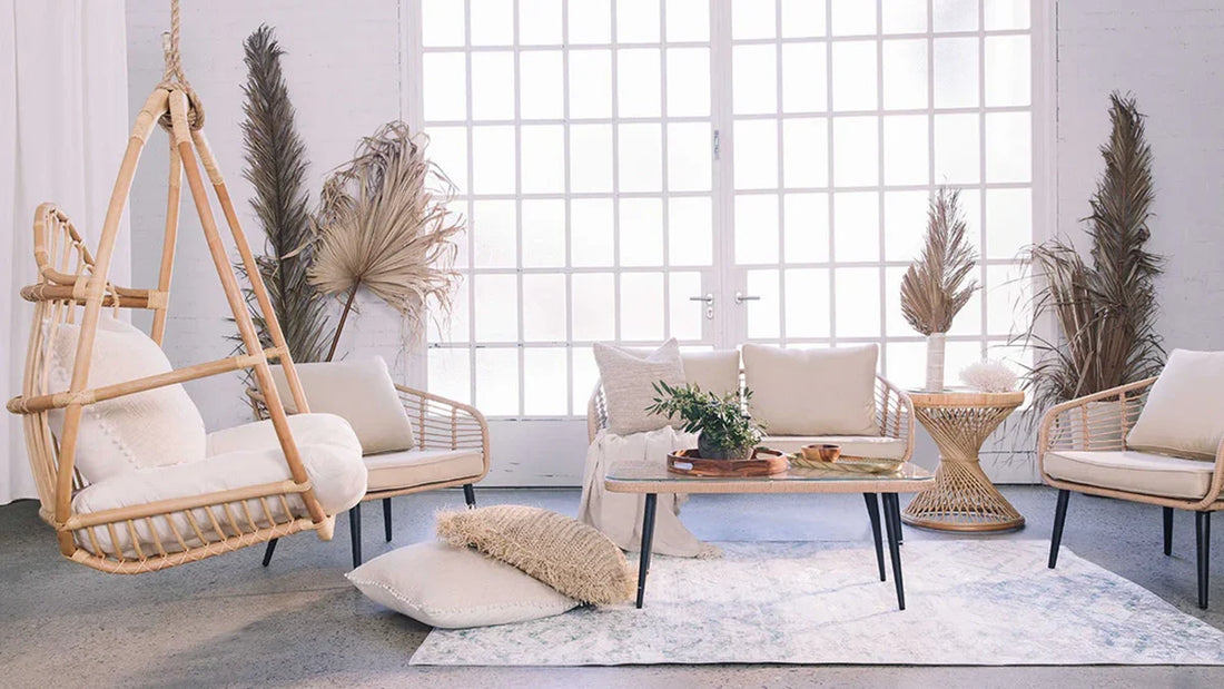 Boho-style living room with a woven rattan hanging chair, matching armchairs, a two-seat sofa, a low coffee table, and tall windows flooding the space with natural light.