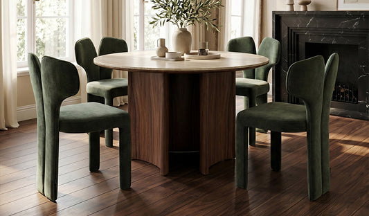 A modern dining set featuring a round, light-toned table with a dark timber, multi-panel pedestal base, surrounded by forest-green, velvet-upholstered dining chairs with a distinctive split-back design, set in a room with a dark marble fireplace.