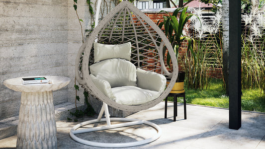 Hilda Large Outdoor Hanging Egg Chair & Cushion in Sage, teardrop-shaped with plush cushions, suspended from a white stand, alongside a small pedestal table and potted plant in a garden setting.