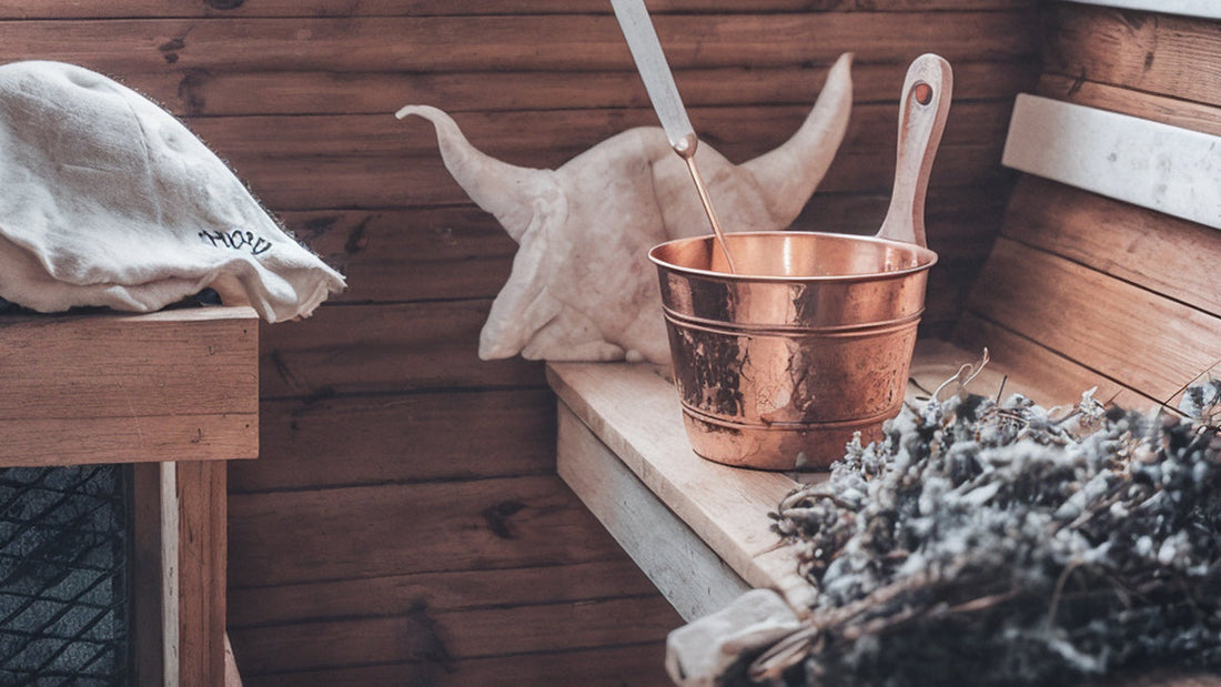 Interior of a wooden traditional sauna with a copper water bucket, ladle, felt sauna hat, and dried lavender for aromatherapy.