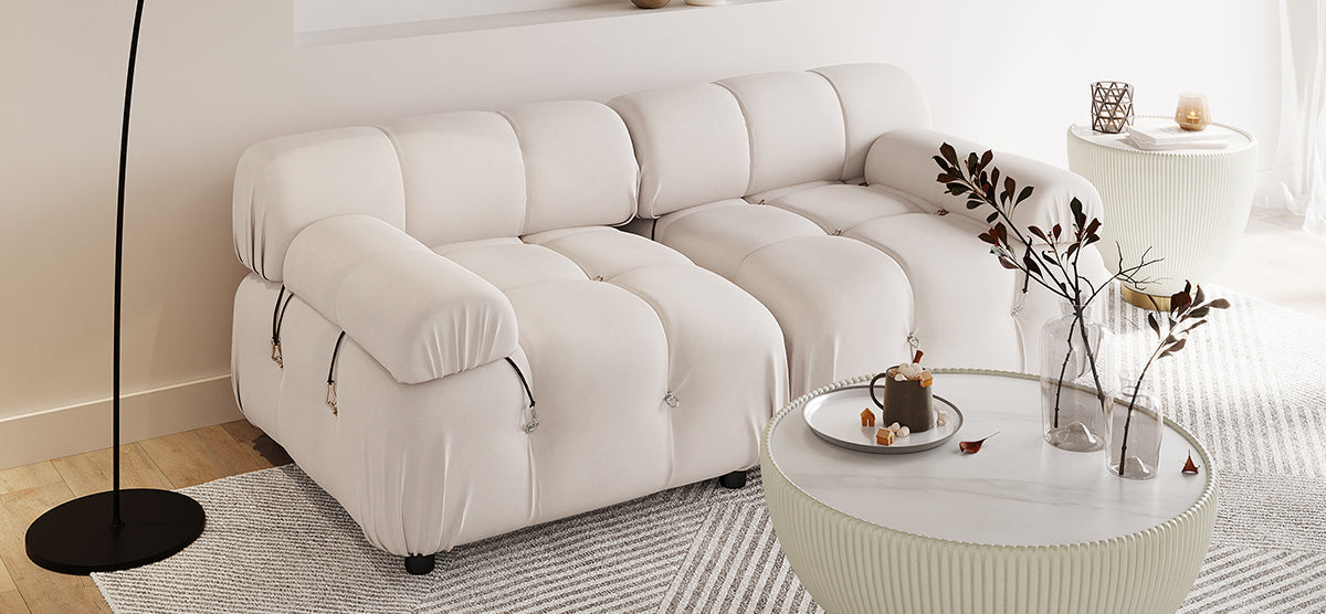 Minimalist living room with a cream tufted sofa, two round fluted coffee tables, glass vases with branches, and a black floor lamp. Neutral rug underfoot.