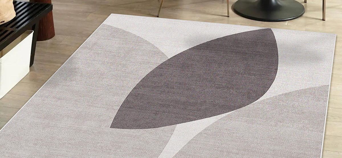 Contemporary area rug with light-gray background, featuring two overlapping leaf motifs in charcoal and pale gray tones, on light wood-tone flooring with partial views of furniture.