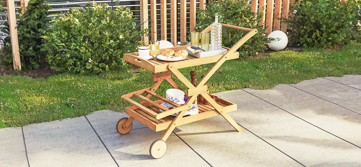 A wooden folding serving cart on wheels, featuring a breakfast setup with croissants and drinks on top, and ceramic dishes below, set on a stone-tiled terrace.