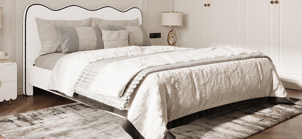 Neutral-toned bedroom with low-profile bed featuring an upholstered headboard, layered linens, and throw pillows. Nearby, a white nightstand holds books and a modern lamp.