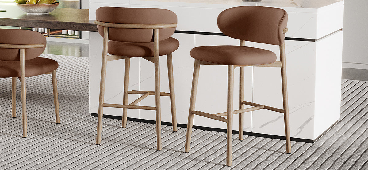 Two counter-height stools with light wood frames and curved brown padded seats are positioned at a white marble kitchen island, adjacent to a dining table with a fruit bowl.