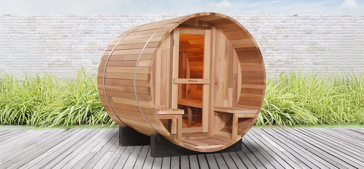 Barrel-shaped wooden sauna with glass-paneled door, two bench seats inside, set on a wooden deck with a low masonry wall and ornamental grasses behind it.