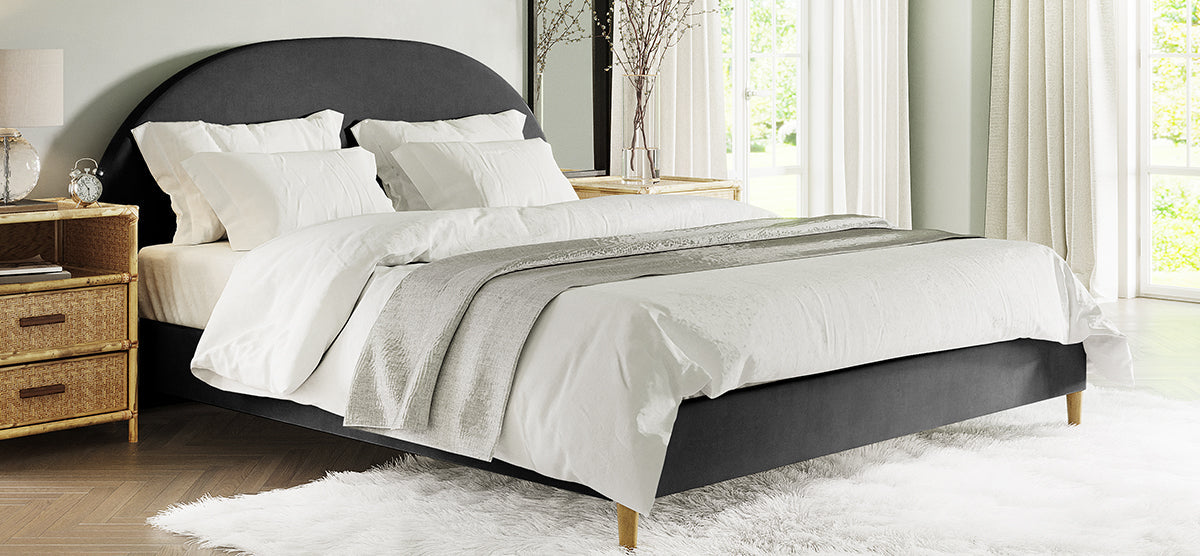 A bright bedroom featuring a dark-gray upholstered bed with white bedding and a gray throw, flanked by rattan tables with a lamp, books, and a mirror.