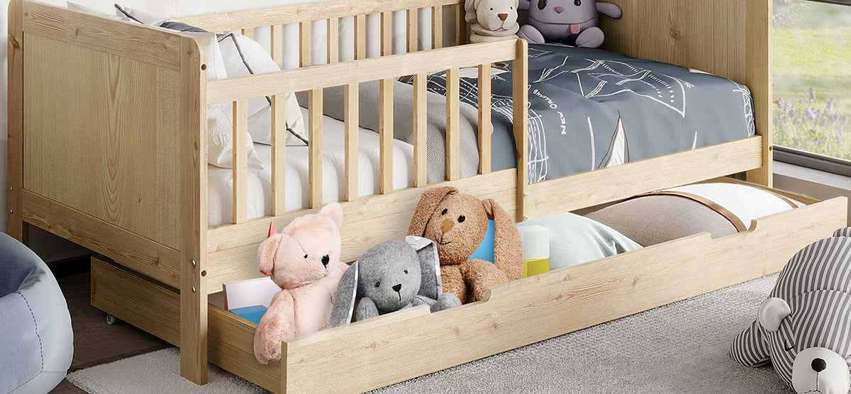 Light-wood children's daybed with pull-out trundle containing plush toys. Top mattress features a white sheet and blue comforter. Nearby, a blue floor cushion and striped stuffed animal.