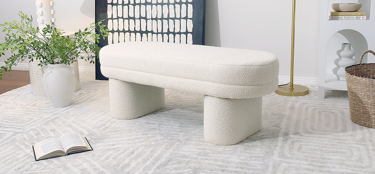 Minimalist living space corner with a cream boucle upholstered bench, geometric rug, ceramic vases with foliage, abstract artwork, brass floor lamp, and a decorative side table.