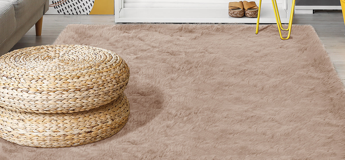 Cozy corner with a beige-pink shag rug, stacked seagrass poufs, gray sofa edge, white shelving with sandals, and a table with bright yellow metal legs.