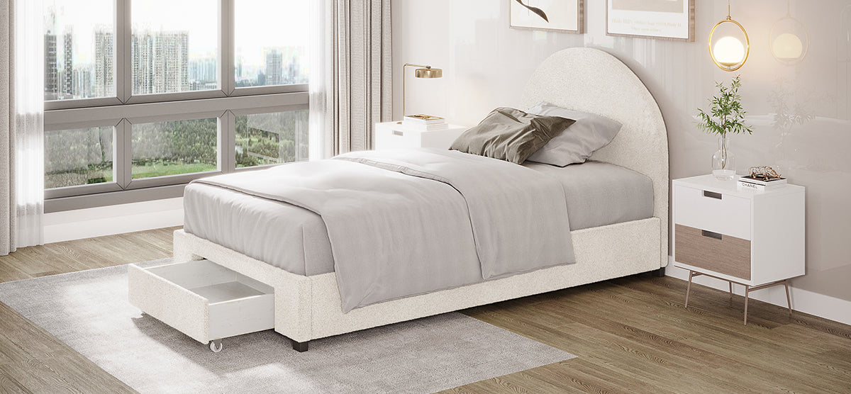 Modern bedroom corner with cream upholstered bed, light gray bedding, under-bed storage, white nightstand with decor, brass lamp, and large windows showing urban skyline and park.