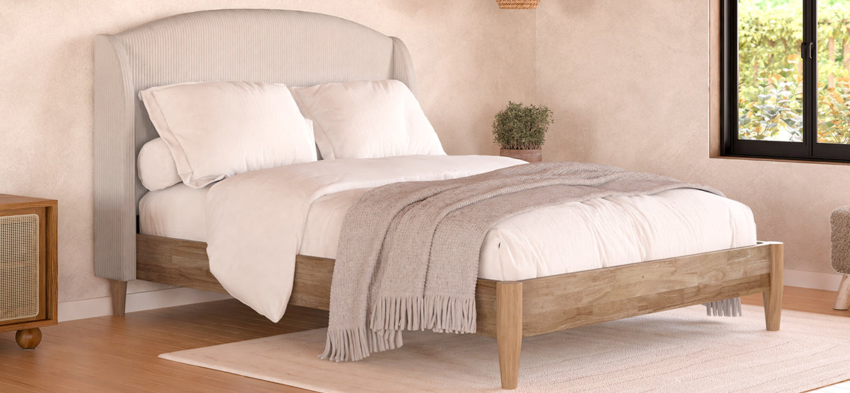 A softly lit bedroom with a wooden-framed bed, light gray headboard, white bedding, taupe throw, wooden nightstand with plant, sandy beige walls, and a window with greenery.