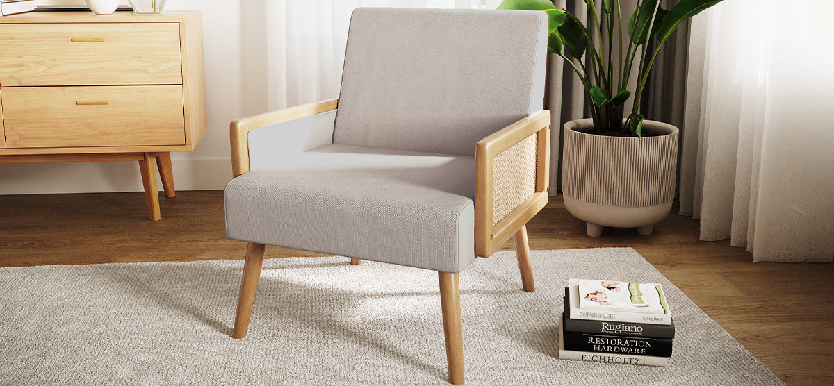 A cozy interior features a light gray armchair with natural wood accents, a wooden sideboard, a ribbed ceramic planter with a leafy plant, and hardcover books on a light rug.