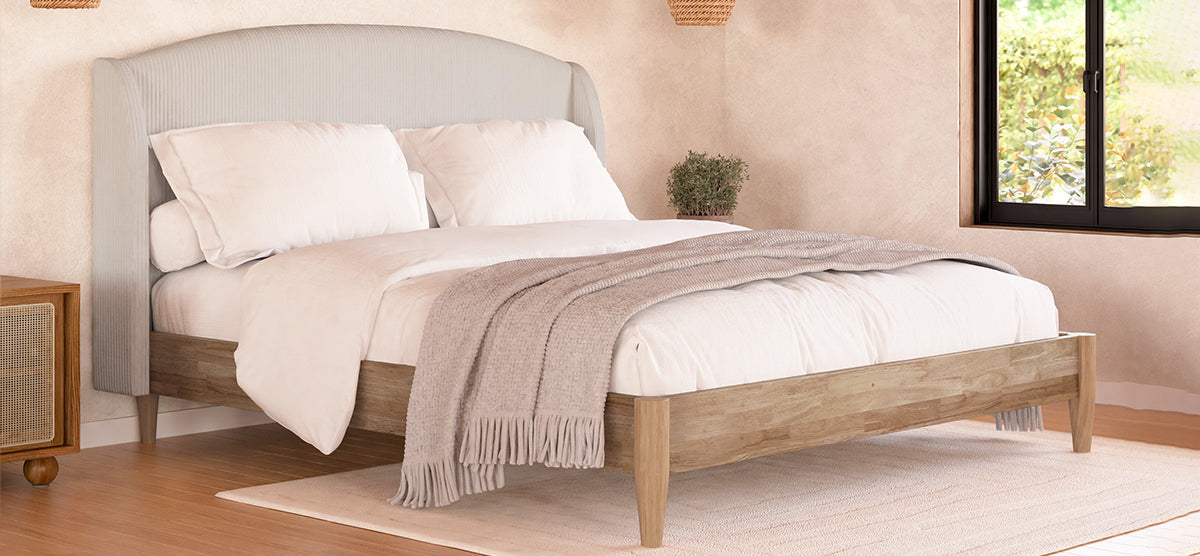 Low-profile wooden bed with light upholstered headboard, white sheets, fringed throw, on pale rug. Nearby, a cane-detailed nightstand and potted plant; window reveals sunlit greenery.