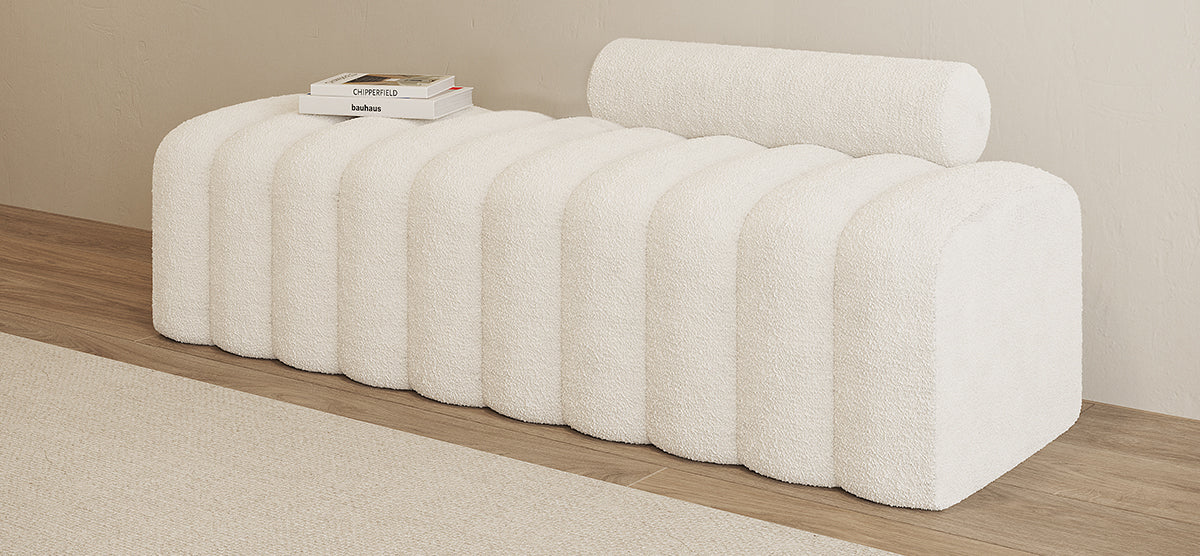 A sculptural bench upholstered in white bouclé with rib-like segments and a cylindrical bolster cushion, accompanied by books titled Chipperfield and bauhaus on a light wood floor.