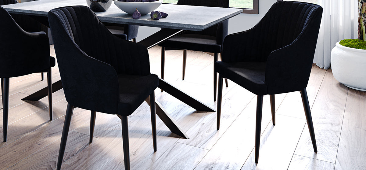 Modern dining area with a stone tabletop, four black upholstered chairs, light wood floor, decorative figs on table, large white planter with plant, and softly draped window.
