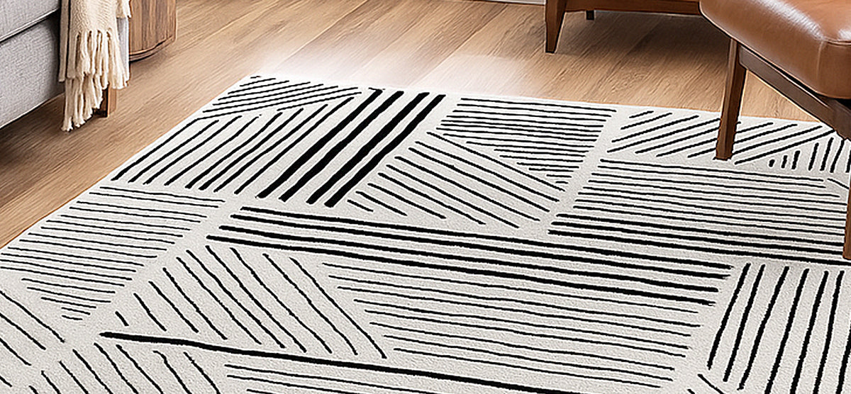 Light-colored area rug with a geometric pattern of black lines on a hardwood floor, alongside legs of an upholstered chair draped with a throw and a wooden side table.