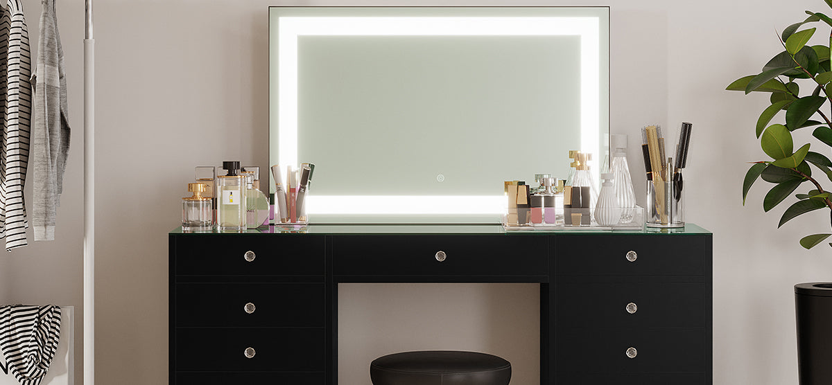 Modern dressing area with a black vanity table, backlit mirror, arranged beauty items, black cushioned stool, potted plant, and garment rack. Ideal for grooming or makeup application.