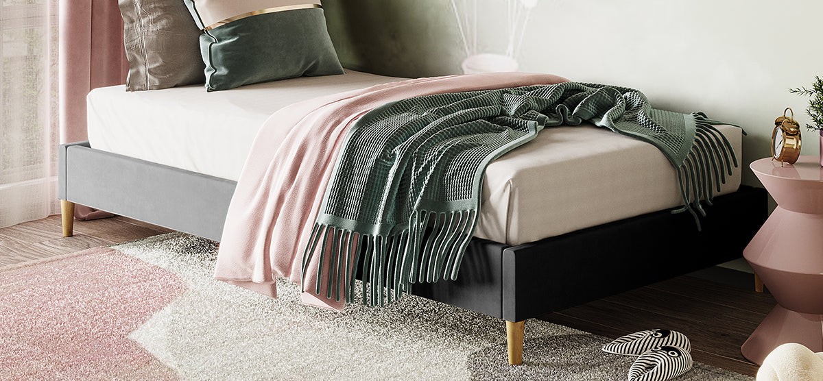 Cozy bedroom corner featuring a slate gray upholstered bed frame with a white mattress, dusty pink and sage green throw blankets, and accent pillows. A pink side table with a plant and gold-trimmed alarm clock.