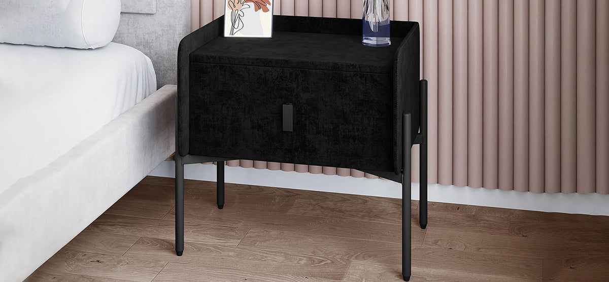 Modern bedroom with a black nightstand featuring slender legs, topped with a framed art print and tall glass vase, beside a white upholstered bed.