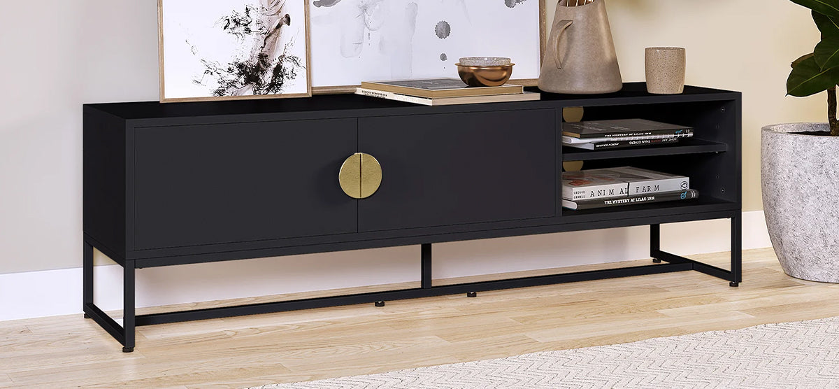 Matte black media cabinet with circular brass pull, slender metal base, open shelves with books, framed prints, decorative items, and a large potted plant beside it.