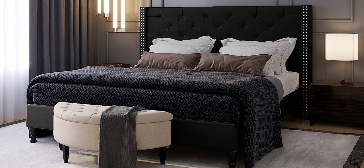 Modern bedroom with a tall, tufted black headboard, white and brown pillows, dark bedspread, upholstered bench with throw, charcoal drapes, bedside tables, and stylish lighting.
