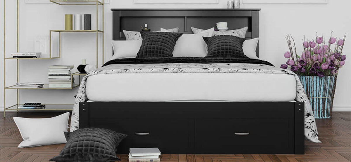 Modern bedroom with a black wooden platform bed featuring built-in storage, patterned duvet, and assorted pillows. Gold shelving unit and woven basket with tulips add decorative touches.