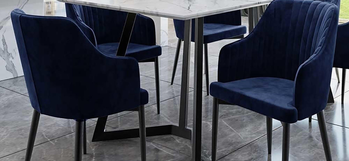 Modern dining setup featuring a rectangular white marble-effect table with black metal legs, surrounded by navy velvet upholstered chairs with curved backs and armrests on a gray tiled floor.