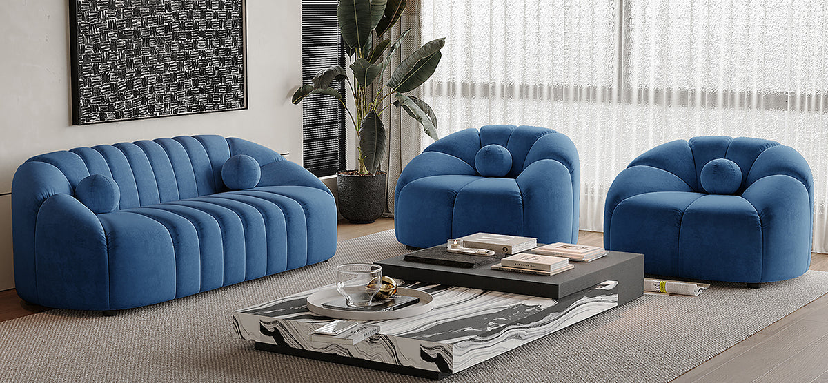 Blue velvet seating group with three-seat sofa and two rounded armchairs, complemented by a two-tier coffee table, textured rug, potted plant, abstract print, and sheer curtains.