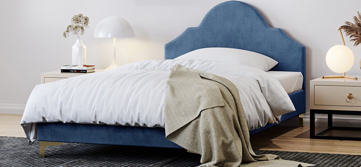 Modern bedroom with a low-profile bed featuring a blue upholstered headboard, white sheets, tan throw, and matching nightstands with decorative lamps, books, and dried flowers.