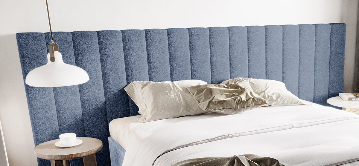 Modern bedroom with a tall, blue-gray upholstered headboard. Features white duvet, matching pillows, two round bedside tables, a teacup, books, and a white pendant lamp.