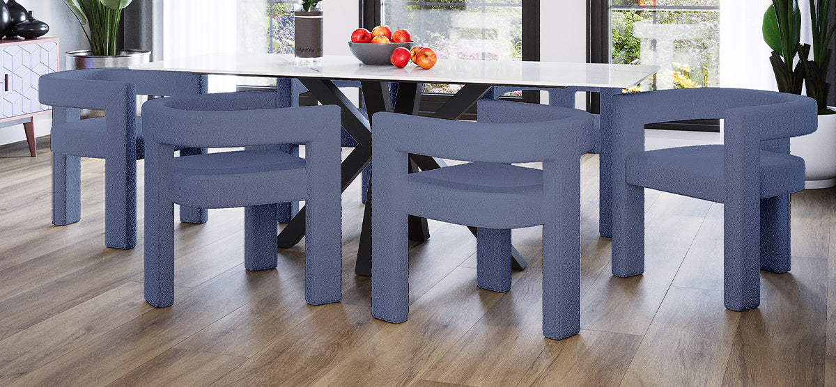 Modern dining area with a white-top table on black X-shaped legs, surrounded by six muted blue, low-back chairs. Features wood-look flooring, large windows, and a fruit bowl centerpiece.