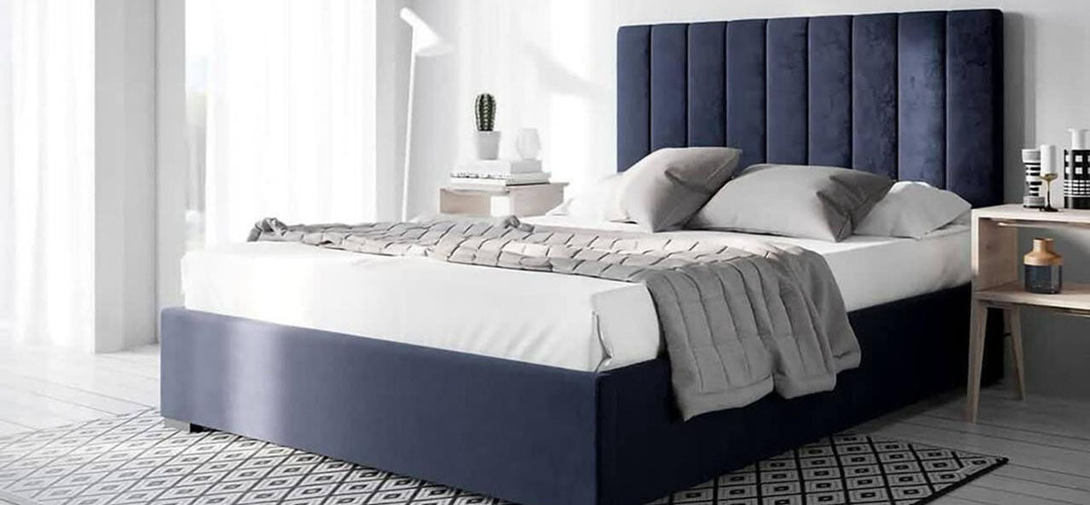 Modern bedroom with navy blue bed featuring a tall, channel-stitched headboard, white and gray bedding. Light wood nightstands, white lamp, books, cactus, decorative vessels, diamond-patterned rug, sheer curtains.