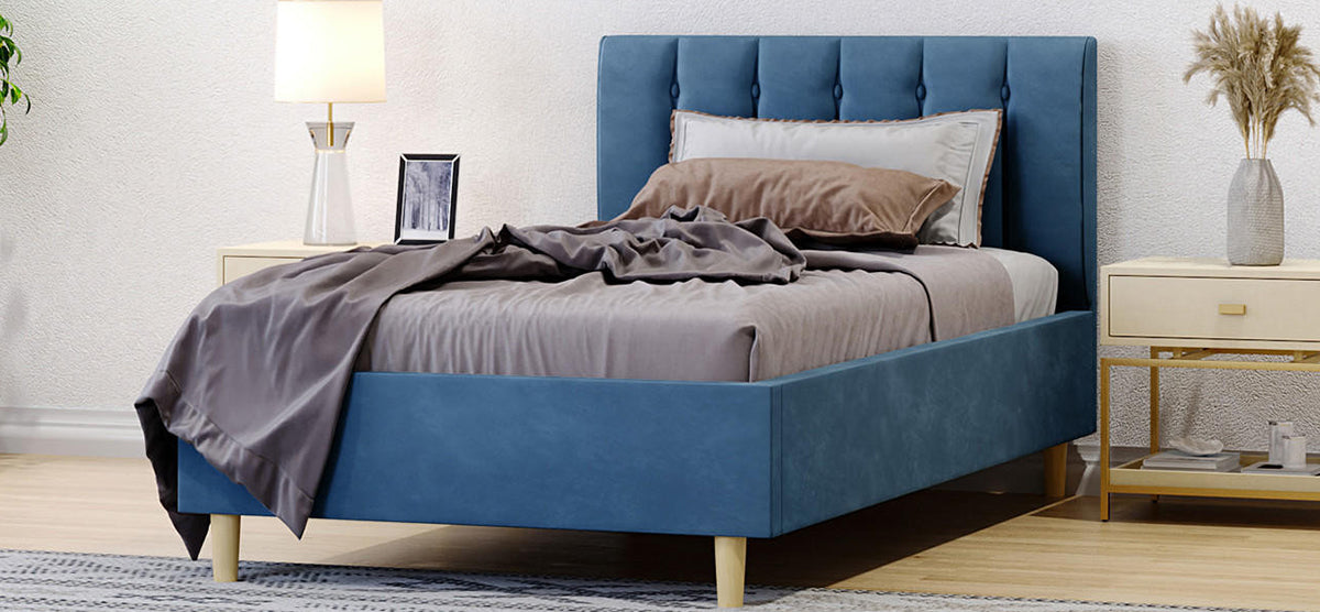 Modern bedroom with a muted blue upholstered platform bed and button-tufted headboard, flanked by light wood nightstands, featuring a lamp, photo, vase with pampas grass, and books.