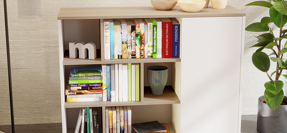 Light-wood shelving unit with cubby shelves filled with books, a decorative m, striped vase, and topped with wooden bowls; flanked by a rubber plant and a floor lamp.