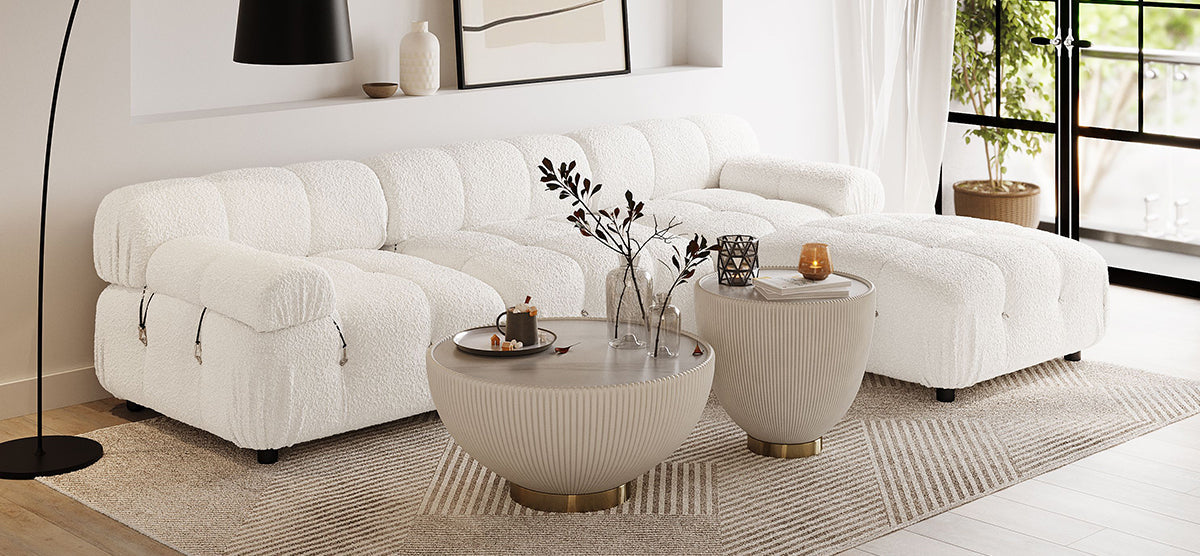 A Scandinavian-inspired living room corner with a white bouclé sectional sofa, sand-tone coffee tables, a black arc lamp, and a patterned rug under a large window.